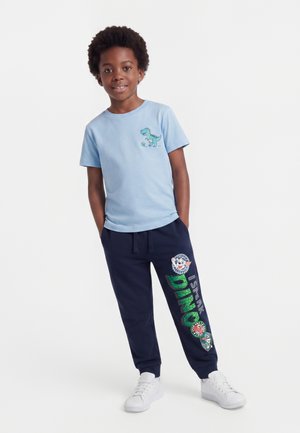 Enfant portant un t-shirt bleu clair avec un motif de dinosaure et un jogging bleu marine avec des patchs colorés sur le thème des dinosaures et des baskets blanches.