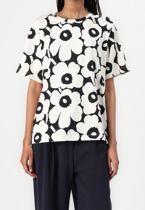 Persoon draagt een zwart-wit bloemenpatroon kortemouwen overhemd met grote bloemen en effen donkere broek.