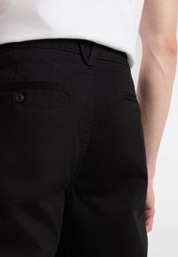 Pantaloni in cotone neri con vestibilità slim, dotati di due tasche posteriori e un dettaglio con un bottone, mostrati da dietro e di lato.