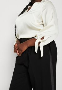 Weiße strukturierte Bluse mit gebundenen Ärmeln kombiniert mit hochtaillierten schwarzen Hosen, die mit einer seitlichen Streifen-Detail versehen sind.