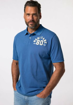 Mand med skæg iført blå Heritage 80 polo og lyseblå jeans, stående med hænderne i lommerne mod en ensfarvet baggrund.