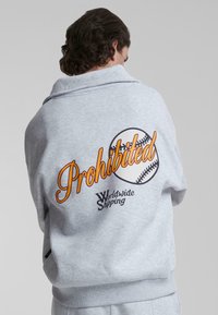 Grå hettegenser med en avslappet passform, med "Prohibited" i oransje skrift, et baseball-illustrasjon, og teksten "Worldwide Shipping" under.