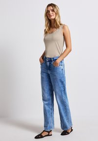 Top a canotta beige, jeans a palazzo blu chiaro, scarpe nere basse. Modella in piedi con le mani in tasca, capelli che cadono sulle spalle.