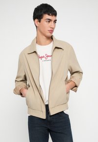 Mladý muž s krátkymi tmavými vlasmi, oblečený v béžovej bunde na zips cez biele tričko Pepe Jeans London a tmavé džínsy, s rukami v kapsách, pozerá doprava.