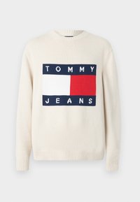 Maglione color crema con polsini e scollatura a costine. Presenta un logo grafico in blu navy, bianco e rosso con la scritta “TOMMY JEANS” centrata sul davanti.