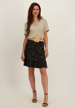 Vrouw staat en draagt een beige V-hals top met korte mouwen, een zwarte bloemenrok met ruches aan de zoom, een brede geweven riem en zwarte enkellaarsjes met hak.