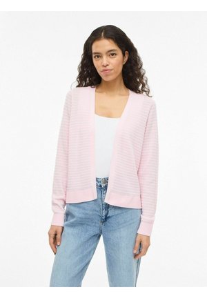 Mujer con cabello rizado lleva un cárdigan rosa claro texturizado sobre una camiseta blanca y jeans azul claro, de pie contra un fondo blanco.