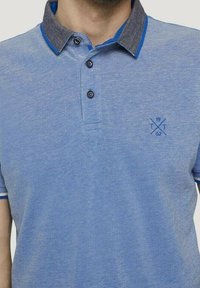 Polotröja i ljusblått med en mörkgrå krage, med en blå detalj, tre knappar och en logotyp på bröstet. Strukturerad tyg.