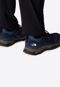 Siniset vaelluskengät, joissa on verkkomateriaali yläosassa, kumipohja ja Vibram-logo. Mustat nauhat ja North Face -logo sivulla.