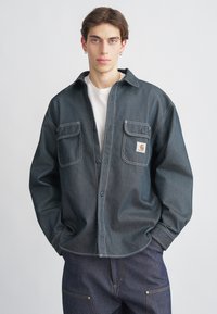 Tmavě modrá džínová košile na knoflíky s dvěma náprsními kapsami a kontrastním bílým prošitím. Nasažená přes bílé tričko, model pózuje před jednobarevným pozadím.