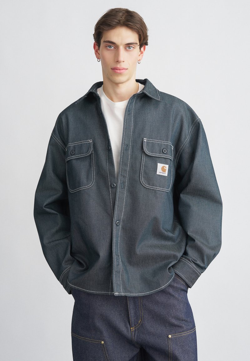 Tmavě modrá džínová košile na knoflíky s dvěma náprsními kapsami a kontrastním bílým prošitím. Nasažená přes bílé tričko, model pózuje před jednobarevným pozadím.