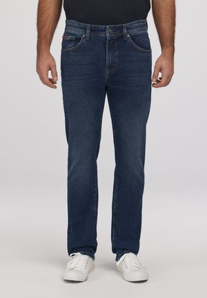 Mężczyzna ubrany w ciemnoniebieskie jeansy o prostych nogawkach i białe trampki, stojący na tle jednolitego jasnego tła.