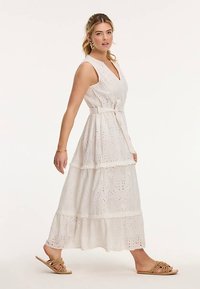 Biała, bezrękawnikowa sukienka z falbanami, zdobiona haftem dziurkowanym i paskiem w talii. Noszona z płaskimi sandałami, które mają pleciony wzór na paskach.