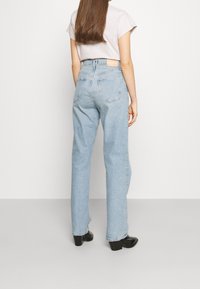 Pitkähiuksinen ruskeatukkainen nainen, joka pukeutunut vaaleansinisiin korkeavyötäröisiin suoralahkeisiin farkkuihin, vaaleanharmaaseen lyhythihaiseen paitaan ja mustiin korollisiin saappaisiin.