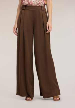 Donna che indossa pantaloni marroni a vita alta e gamba ampia abbinati a sandali aperti e top floreale senza maniche, in piedi su uno sfondo neutro.