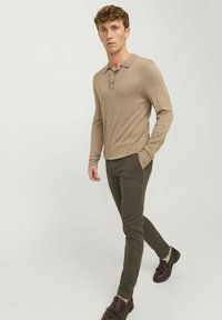 Beige Polo-Shirt mit langen Ärmeln, Kragen und drei Knöpfen, kombiniert mit dunkelgrünen Slim-Fit-Hosen und braunen Schuhen mit strukturiertem Finish.