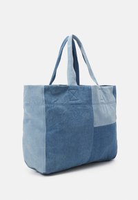 Denim tote taske med et patchworkdesign i varierende nuancer af blå, robust stof, to lange håndtag og en firkantet bund.