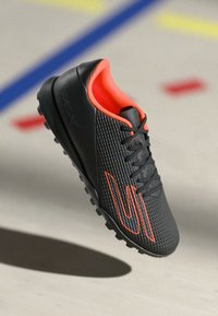 Black textured sports shoe with orange inner lining, orange logo, black laces, and rubber sole with small cleats on indoor court.