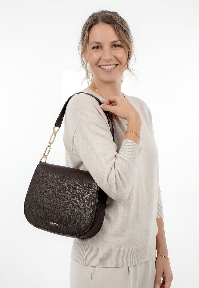 Donna sorridente con maglione e pantaloni beige, che tiene una borsa a tracolla in pelle marrone scuro con dettagli dorati.