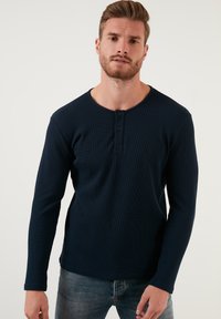Mornarsko majico s dolgimi rokavi henley iz strukturirane tkanine, ki ima okroglo izrez in tri gumbe, v kombinaciji z jeansa.