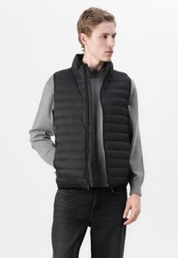 Zwarte puffer vest met een hoge kraag, gewatteerde textuur, ritsluiting en een klein merklogo op de borst, gedragen over een grijze trui.