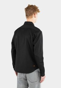 Svart langermet skjorte med strukturert krage og knappeender. Har en avrundet hem og små oransje merke detaljer nær bunnen.