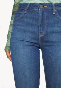 Gros plan de la partie centrale portant un jean bleu Lee avec coutures visibles, bouton et poches avant, associé à un haut vert à manches longues.