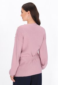 Dammrosa stickad kofta med långa ärmar, ribbat mönster, bälte i midjan och pärldetaljer på axlarna. Visad från baksidan.