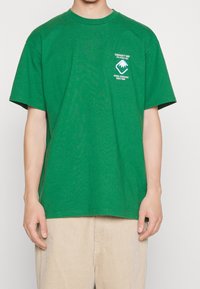 Camiseta de algodón verde con mangas cortas, cuello redondo y gráficos impresos en blanco en el lado izquierdo del pecho y la parte trasera. Combinada con pantalones de pana beige.