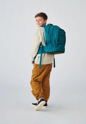 Teal backpack with padded straps, multiple compartments, and side pockets. Worn by a child in a white sweater and cargo pants.