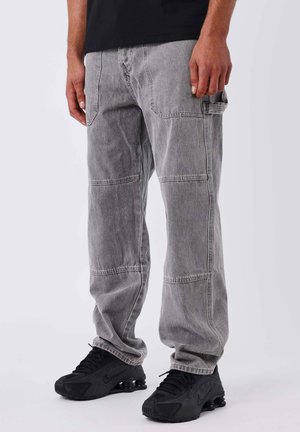 Man wearing faded gray cargo jeans and black Nike sneakers, standing against a plain white background.