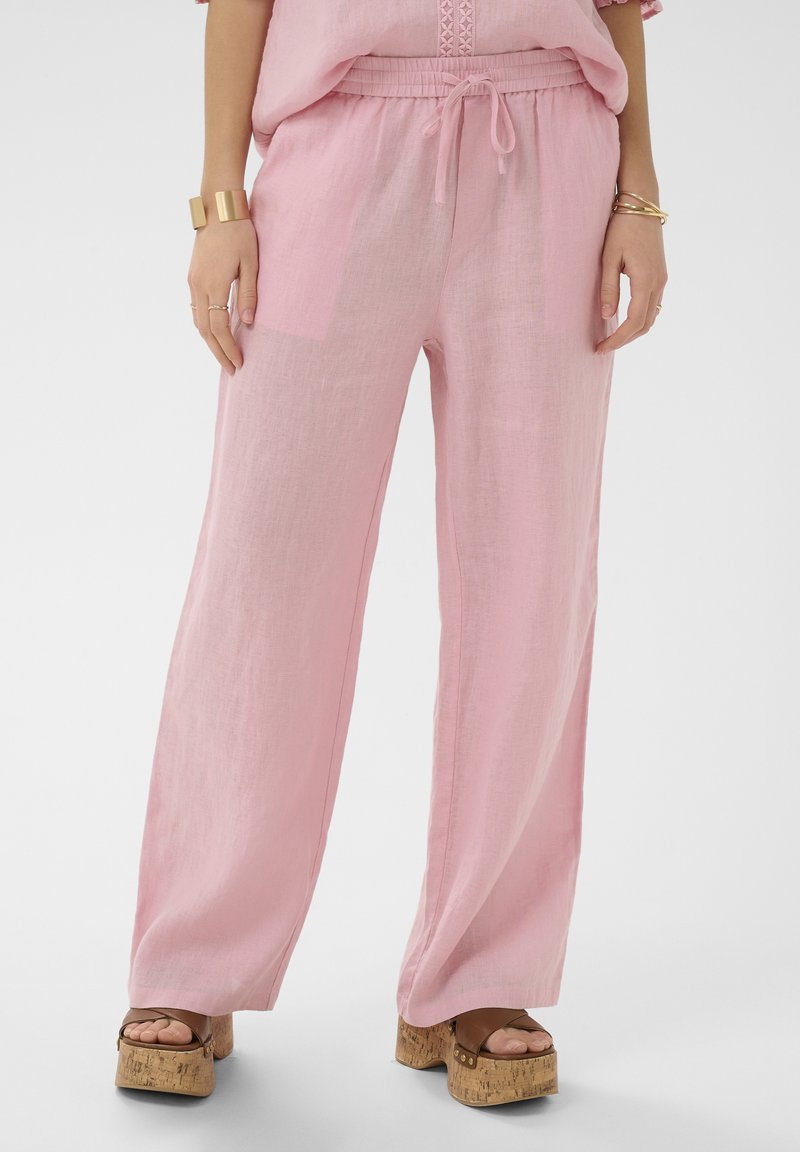 Femme portant un pantalon ample en lin rose avec une taille élastique à cordon de serrage et des sandales à plateforme en liège, montrant le bas du torse et les jambes.