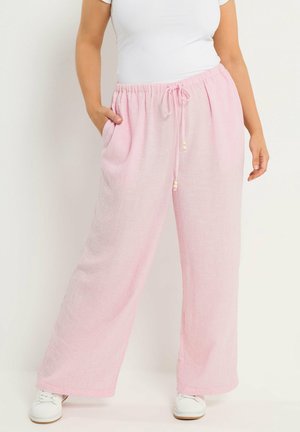 Frau trägt lockere, hellrosa Hosen mit Kordelzug und weiße Turnschuhe, eine Hand in der Tasche, vor hellem, einfarbigem Hintergrund.