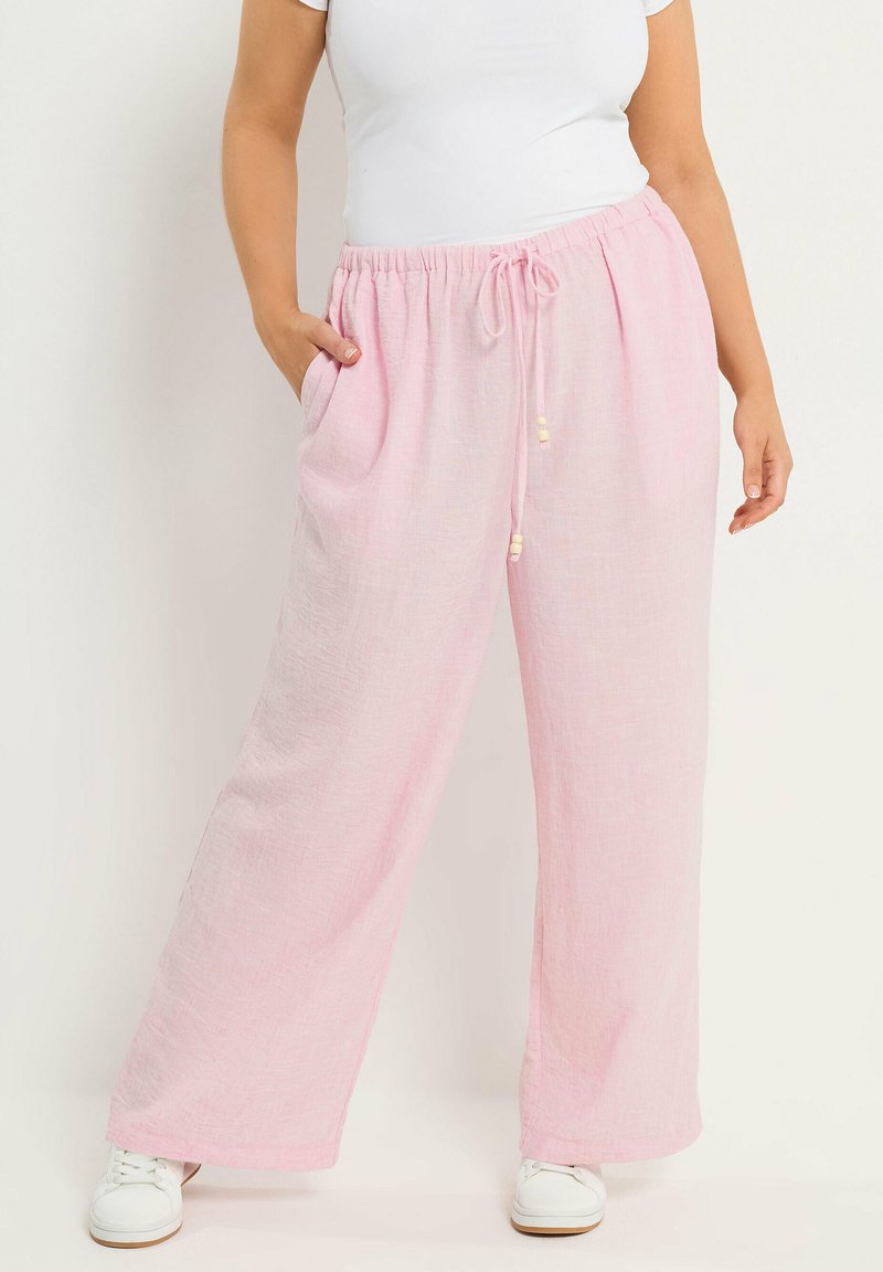 Frau trägt lockere, hellrosa Hosen mit Kordelzug und weiße Turnschuhe, eine Hand in der Tasche, vor hellem, einfarbigem Hintergrund.