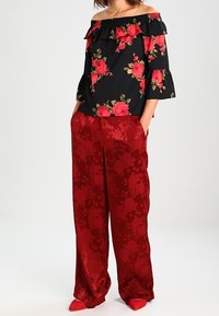 Woman wearing black off-shoulder top with red floral print, paired with wide-leg red satin pants with floral texture and red pointed shoes.