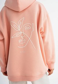 Hoodie rosa con un outline ricamato bianco di un volto e foglie sul retro. Il materiale è morbido, con una vestibilità comoda e cappuccio.