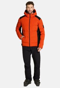 Doudoune orange vif avec un design à panneaux noirs, fermeture éclair à l'avant et capuche. Associée à un pantalon noir et des bottes de ski. Texture lisse.
