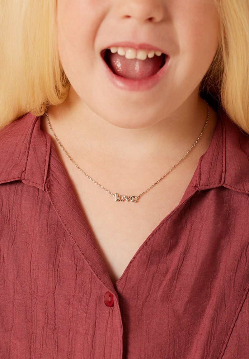 Gouden halsketting met een "love" hanger, gemaakt van metaal, gedragen over een maroon textuur blouse met kraag en enkele knoopdetail.