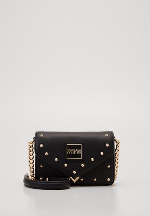 Zwarte Versace Jeans Couture crossbody tas met gouden studs, logoplaatje en een gouden kettingriem, rustend op een effen lichte achtergrond.
