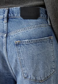 Ljust blå denimjeans med en texturerad finish, som har en svart läderlapp med texten "ALLSAINTS" på midjebandet och en bakficka.