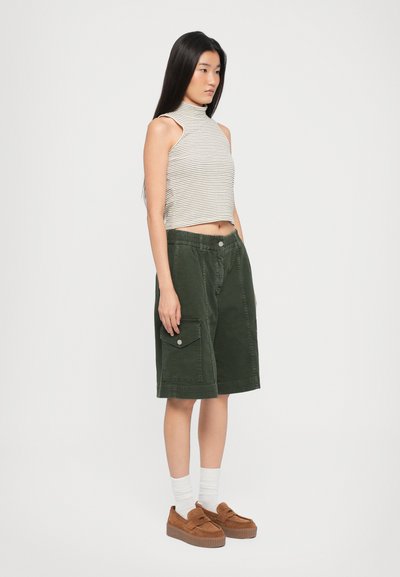 Λωρίδες χωρίς μανίκια τοπ, πράσινες βαμβακερές βερμούδες με πλαϊνές τσέπες, καφέ υφασμάτινα παπούτσια με ρόκα, φορεμένα με λευκές κάλτσες, στέκονται μπροστά σε ένα απλό �φόντο.