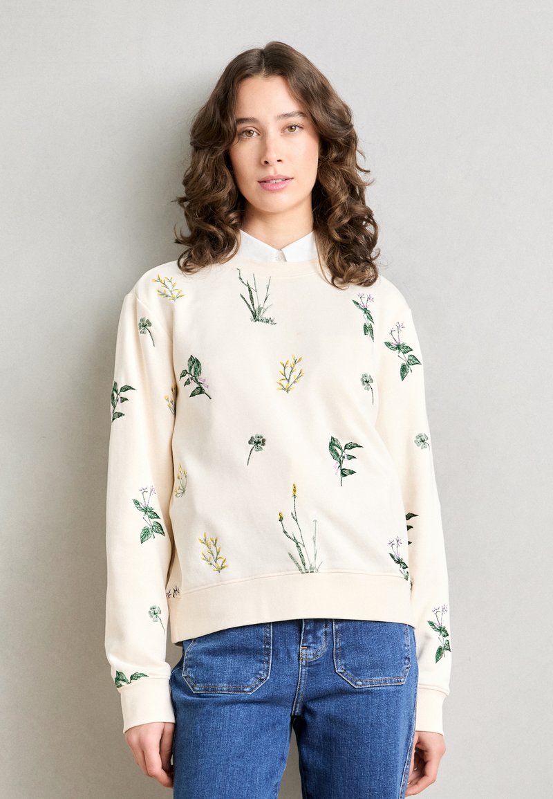 Krem gensere med blomsterbroderi i grønne og gule nyanser. Har rund hals og ribbede mansjetter. Kombinert med blå denimjeans.