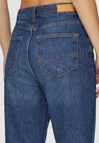Donkerblauwe denim jeans hebben een hoge taille, strakke lijnen, twee achterzakken en een tan leren patch op de tailleband.