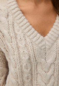 Ljusbeige stickad tröja med djupt V-hals. Har texturerade kabelmönster, ribbade ärmslut och mjukt, lurvigt garn.