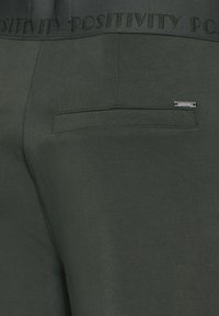 Donkergroene broek met een gladde textuur, voorzien van een enkele achterzak en een tailleband die is gebrandmerkt met "POSITIVITEIT." Zilverkleurige hardware details aanwezig.
