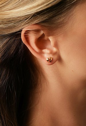 Primo piano dell'orecchio di una donna che indossa un piccolo orecchino a forma di cuore in oro, con capelli biondi e castani visibili.