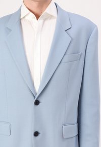 Blazer azul claro con un diseño estructurado, que presenta solapas de muesca, un bolsillo en el pecho y dos bolsillos frontales con botones negros.