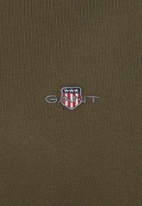 Olive green fabric with a ribbed texture featuring an embroidered GANT logo above a shield with red, white, and blue accents.