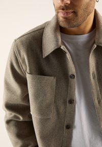 Grey wool jacket with a collared neckline, a chest pocket, and button closures. Features a textured finish and layered over a white shirt.