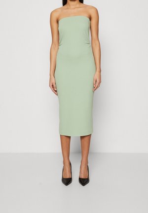 Kobieta ubrana w bezrękawnikową, bezramienną sukienkę midi w jasnym zielonym kolorze, zestawioną z czarnymi, spiczastymi szpilkami, stojąca na tle jednolitego tła.